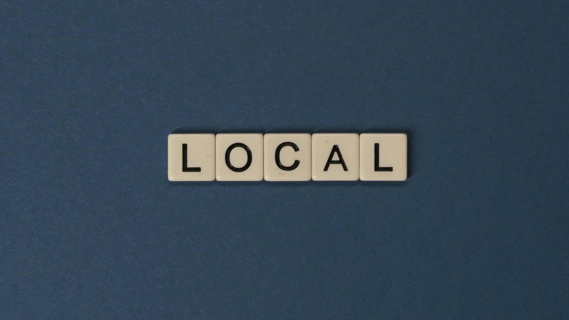 Beige Scrabble tiles spelling LOCAL centered on a solid dark grey'ish blue background.
