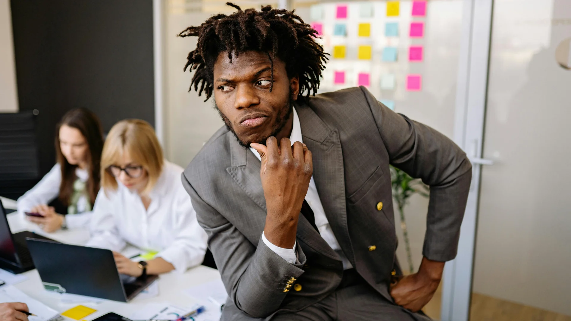 A black man in a gray suit sits in an office conference room, hand on chin in a thoughtful pose, looking sideways with a skeptical expression. Two women work at a desk in the background near a glass wall covered in colorful sticky notes.